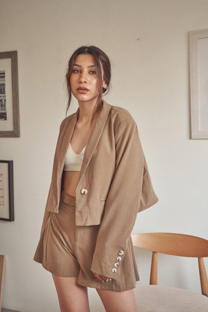 Nadya Inzunza is shown in a room with framed artworks on the wall wearing a beige blazer and matching shorts with a white inner garment visible at the top.